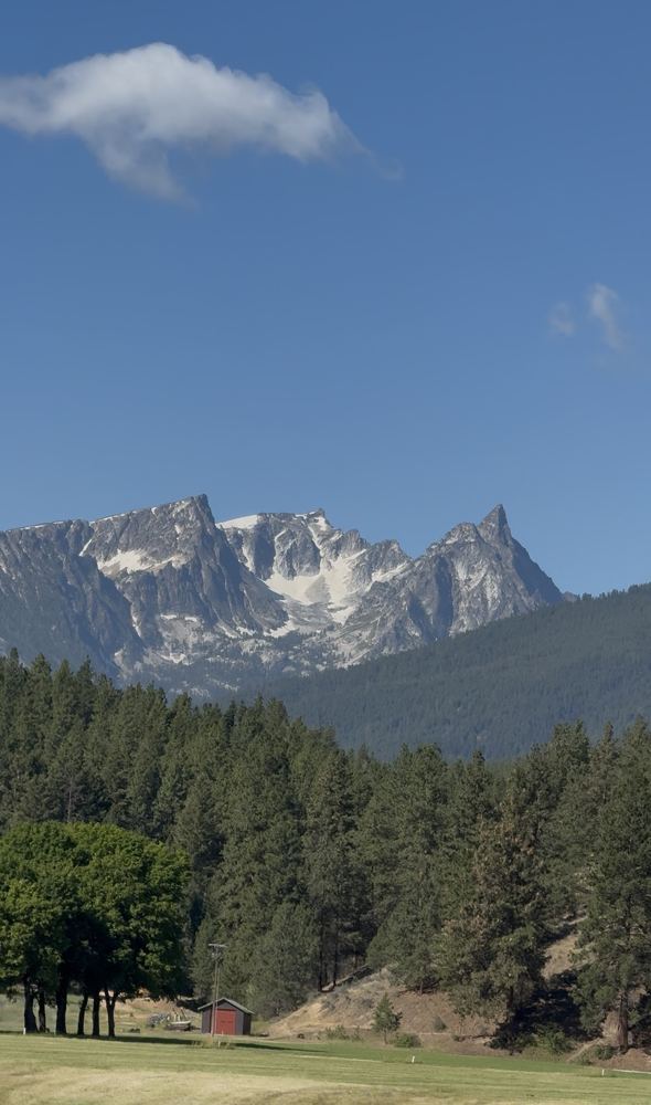 Snow on jagged peaks above a pine forest and a small red barn